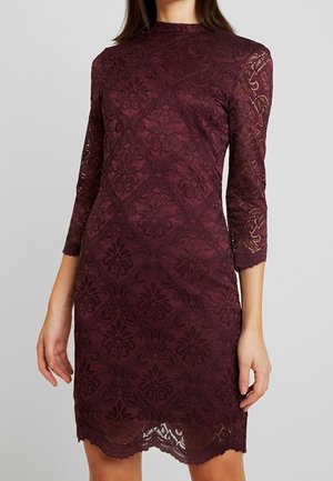 Kobieta ubrana w dopasowaną, do kolan, ciemnofioletową koronkową sukienkę z rękawami za łokieć i kwiatowymi wzorami na neutralnym tle.