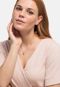Goldfarbener Halsumfang mit rundem Anhänger, getragen über einem hellrosa strukturierten Oberteil mit V-Ausschnitt. Glatte Haut und sommersprossiger Teint.