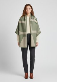 Poncho en mélange de laine en vert olive et beige avec un motif géométrique ; tombe sur les épaules ; associé à un pantalon gris et des bottes marron.