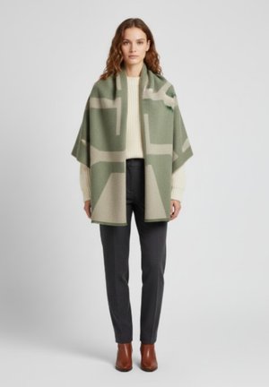 Poncho en mélange de laine en vert olive et beige avec un motif géométrique ; tombe sur les épaules ; associé à un pantalon gris et des bottes marron.