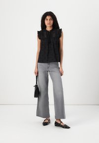 Top nero senza maniche con spalline arricciate, ritagli decorativi e dettaglio a fiocco. Abbinato a jeans grigi a gamba larga e scarpe nere flat.