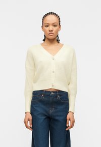 Femme aux cheveux tressés portant un cardigan en maille crème boutonné et un jean taille haute bleu foncé, debout devant un fond blanc uni.