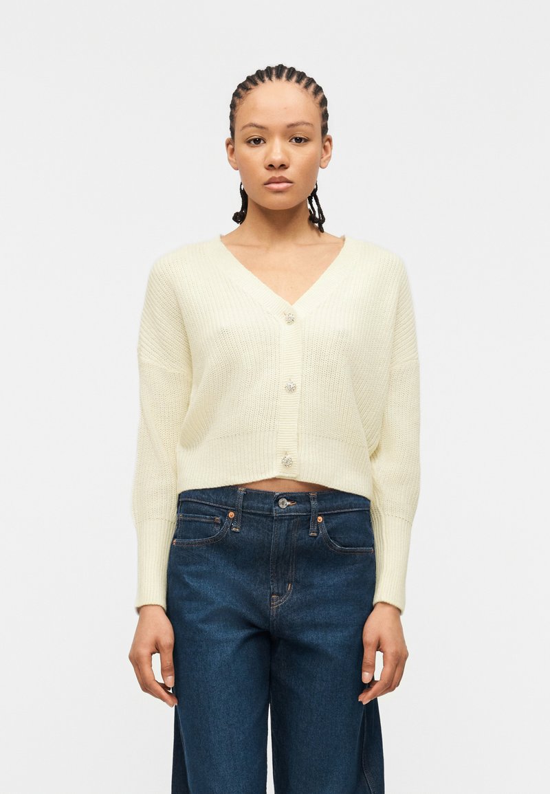 Femme aux cheveux tressés portant un cardigan en maille crème boutonné et un jean taille haute bleu foncé, debout devant un fond blanc uni.