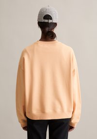 Brzoskwiniowy, oversize'owy sweter z prążkowanym okrągłym dekoltem, opadającymi ramionami i bezszwowymi mankietami, w połączeniu z czapką w czarno-skalę.