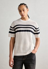 Beiger Strickpullover mit kurzen Ärmeln und schwarzen horizontalen Streifen; Rundhalsausschnitt und strukturierter Stoff, kombiniert mit dunkelblauen Jeans.