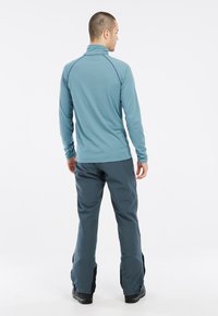 Camicia a maniche lunghe blu chiaro con colletto alto, abbinata a pantaloni impermeabili grigio scuro con zip laterali e una texture liscia.