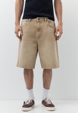 Muž nosiaci béžové voľné džínsové šortky, čierne tielko, biele ponožky a hnedé plátené tenisky stojaci pred jednoduchým pozadím.