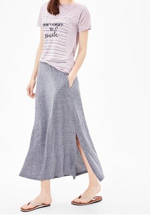 Nainen pukeutuneena lyhythihaiseen vaaleanpunaiseen paitaan, jossa lukee "Älä unohda hymyillä", harmaaseen nilkkapituisen hameen sivuhalkiolla ja beigenväriset varvastossut.