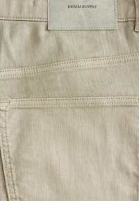 Bež denim tkanina s vidljivim šavovima i kožnom etiketom koja glasi "DENIM SUPPLY." Ima pet džepova i glatku teksturu.