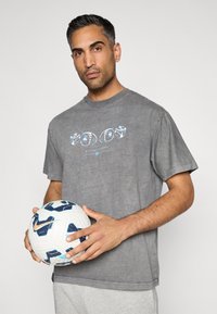 T-shirt cinza de manga curta com uma impressão gráfica com detalhes azuis. O modelo segura uma bola de futebol branca com detalhes azuis e laranjas.