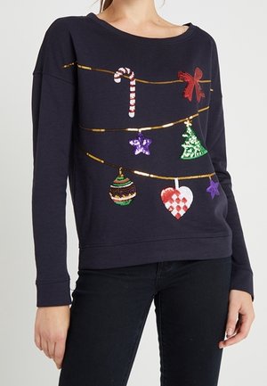 Granatowa bluza z cekinowymi świątecznymi ozdobami, w tym lizakiem, kokardą, gwiazdkami, choinką, bombką i sercem, noszona z czarnymi spodniami.