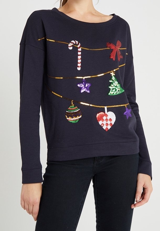 Sweat bleu marine avec décorations de Noël en sequins comprenant sucre d’orge, nœud, étoiles, sapin, boule et cœur, porté avec un pantalon noir.