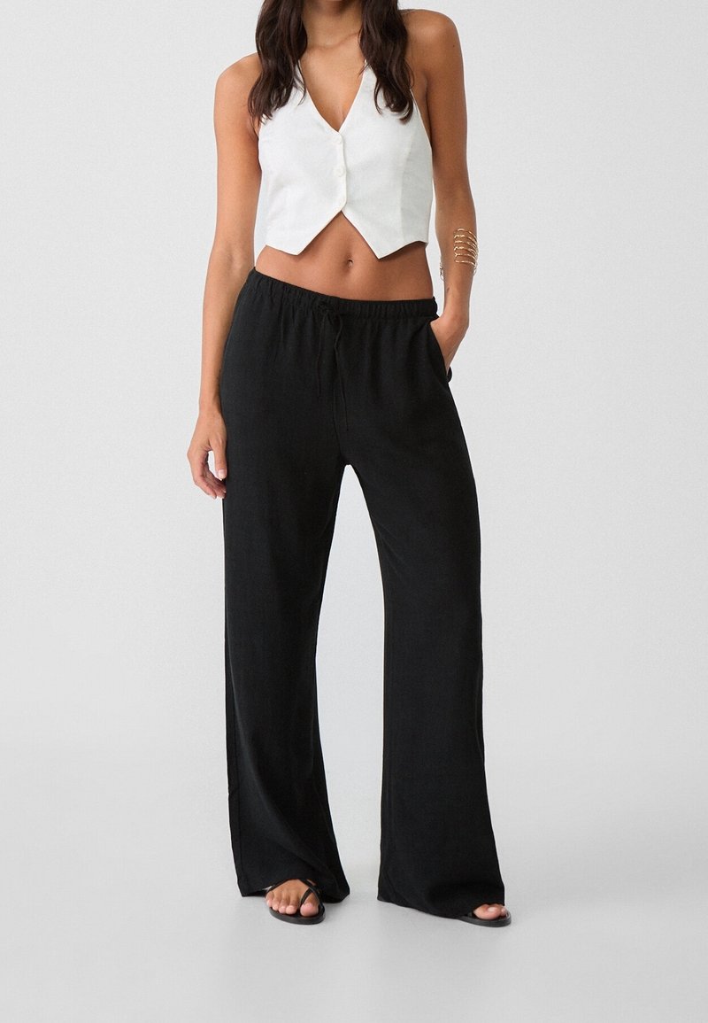 Femme portant un pantalon large noir à taille coulissante, un top court boutonné sans manches blanc et des sandales noires, debout avec une main dans la poche.