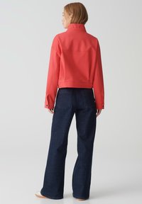 Rote cropped Jacke mit hohem Kragen, aus glattem Stoff gefertigt, kombiniert mit hochgeschnittenen, dunkelblauen Denim-Jeans im klassischen Schnitt und mit Gesäßtaschen.