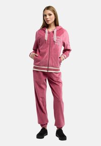 Rosa velourträningsdräkt med zip-up hoodie och elastiska muddar, med vita randiga detaljer och logotyp på bröstet.