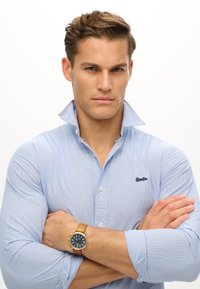 Camicia a righe blu chiaro con un colletto dritto notevole, abbinata a un orologio da polso in tono oro con quadrante blu e cinturino in pelle color cuoio.