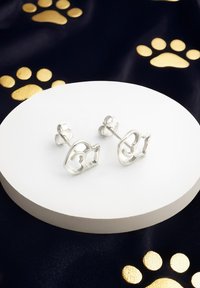 Silver kattformade stiftörhängen med öppen design, som har en slät yta och en polerad finish, placerade på en vit cirkulär pelare.
