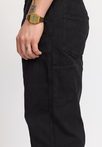 Pantalones cargo negros con múltiples bolsillos, ajuste recto y costuras visibles. Una mano con un reloj dorado descansa en el costado.