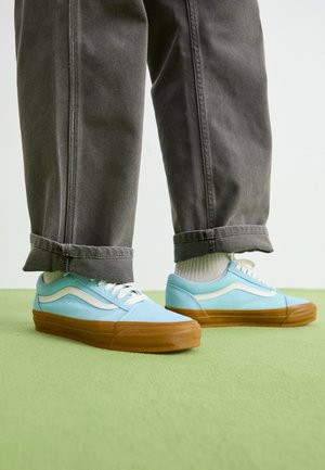 Baskets bleu clair avec des accents blancs, semelles en caoutchouc et lacets. Portées avec un pantalon gris à poignets, présentant un tissu texturé. Surface de sol verte.