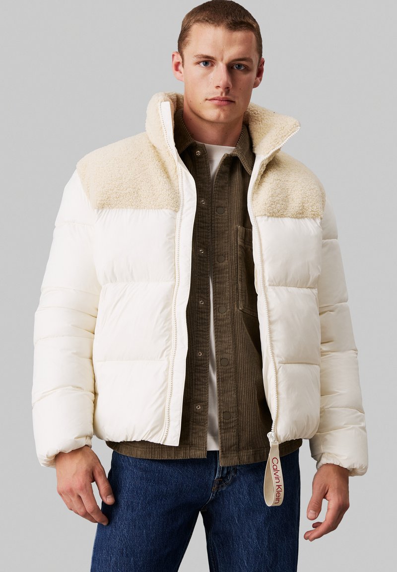 Biała kurtka puchowa z kremowym, sztucznym kołnierzem z owczej wełny, z zamkiem błyskawicznym i metką z logo Calvin Klein, zestawiona z zieloną koszulą z korduroju.