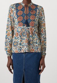 Blusa a fantasia floreale multicolore con scollatura a chiave, dettagli blu e frange. Maniche lunghe e vestibilità comoda.