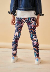 Leggings colorés avec un fond bleu marine ornés de licornes, d'arcs-en-ciel et de motifs floraux. Tissu lisse, associés à des chaussures blanches.