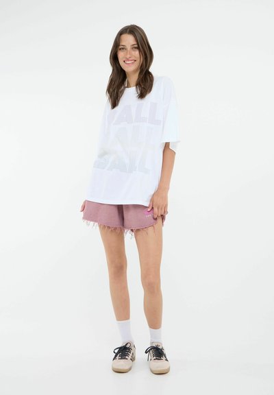 Hvit oversized t-skjorte med "BALL"-tekst i subtile farger, kombinert med lyse rosa slitte shorts. Modellen har på seg svarte og kremfargede sneakers.