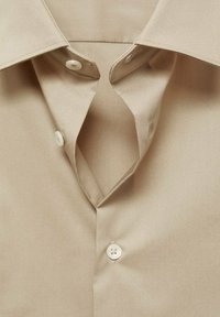 Camicia beige con bottoni a pressione, colletto aperto, bottoni in plastica bianca e tessuto liscio con una leggera lucentezza. Senza motivi o dettagli.