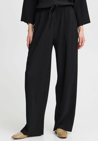 Pantaloni larghi a coste neri con vita elastica e cordoncino. Il tessuto è leggero con una texture liscia. Abbinati a scarpe piatte beige.