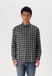 Marmuriniai juodos ir baltos spalvos flaneliniai marškiniai su mygtukų apykakle, ilgomis rankovėmis ir krūtinės logotipo detale; derinami su mėlynomis džinsais.