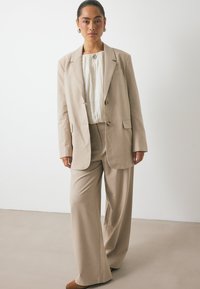 Traje beige a medida con un corte holgado, que incluye un blazer de un solo botón, pantalones de pierna ancha y una blusa texturizada blanca debajo.