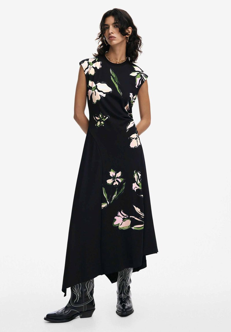 Schwarzes Blumenmaxikleid mit kurzen Ärmeln, das große rosa und grüne Blumen zeigt. Glattes Material mit asymmetrischem Saum, kombiniert mit schwarzen Cowboy-Stiefeln.