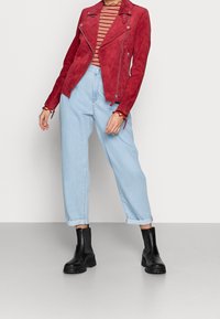 Červená semišová motorkářská bunda se zipy, zkombinovaná se světle modrými zahnulemi denimovými kalhotami a černými kotníkovými botami. Pod tím je pruhované tričko.