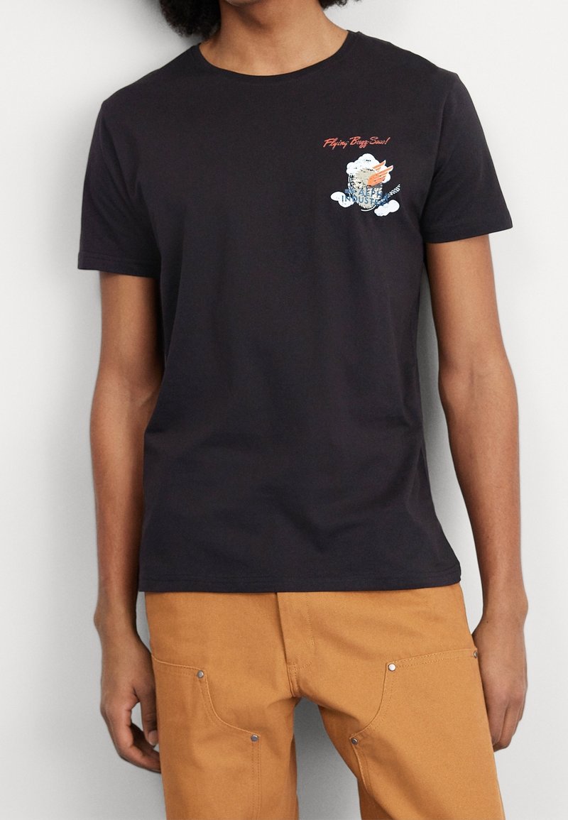 Person wearing a black T-shirt with small orange and white graphic on the left chest and tan pants with front pockets and rivets.