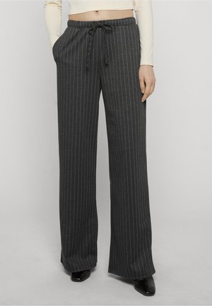 Vrouw die donkergrijze broek met wijde pijpen en krijtstreep draagt, met een trekkoord in de taille en zwarte puntige schoenen, handen in de zakken.