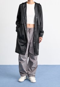 Schwarzer, wasserdichter Trenchcoat mit Kapuze, zwei Vordertaschen; kombiniert mit hellgrauer, locker sitzender Hose und weißen Sneakers.