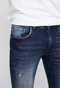 Persona que lleva jeans de mezclilla de color azul oscuro y desgastados, y una camisa blanca, mostrando los bolsillos frontales y laterales con ligeros detalles desgastados.