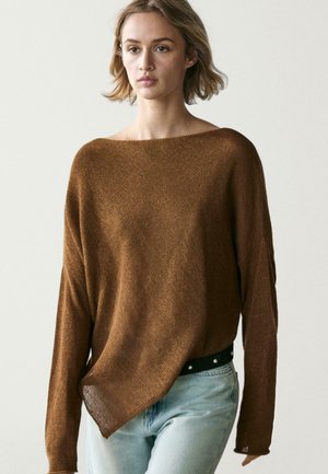 Donna che indossa un maglione marrone lavorato a maglia, ampio, con scollo a barca e jeans azzurri, su uno sfondo chiaro e uniforme.