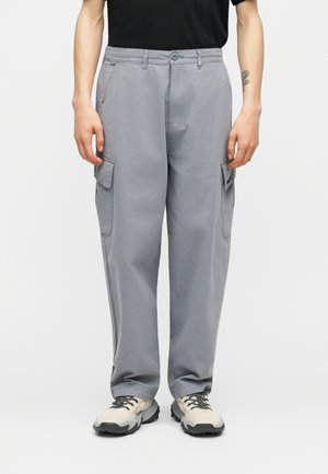 Γκρι φαρδιά παντελόνια cargo με πλαϊνές τσέπες με καπάκια, φορεμένα με μπεζ και γκρι παπούτσια πεζοπορίας και μαύρο πουκάμισο.