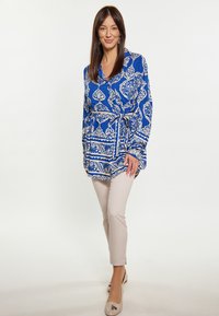 Blusa a maniche lunghe con pattern blu e bianco, vita cintrata, abbinata a pantaloni beige chiaro e scarpe ballerine chiare.