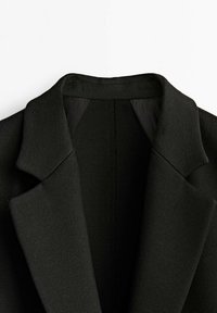 Blazer noir avec un col cranté, en tissu lisse et aux épaules structurées. La doublure intérieure est visible au niveau du cou. Design minimaliste.