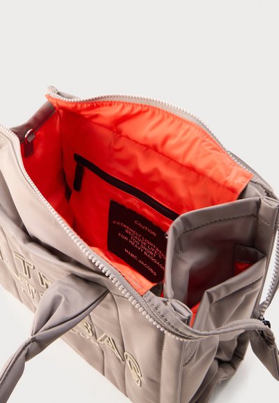 Τσάντα tote σε ανοιχτό γκρι χρώμα Marc Jacobs με πορτοκαλί επένδυση εσωτερικά, κλείσιμο με φερμουάρ επάνω, εσωτερική τσέπη με φερμουάρ και τυπωμένη ετικέτα προειδοποίησης στο εσωτερικό.