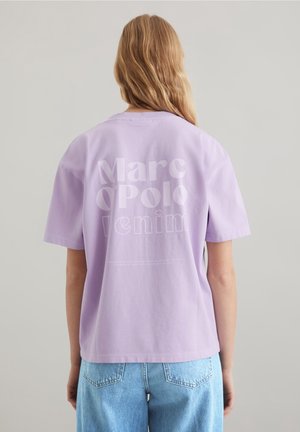 Osoba z długimi blond włosami, ubrana w lawendowy t-shirt Marc O'Polo Denim i jasnoniebieskie dżinsy, zwrócona tyłem na jednolitym tle.