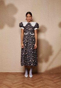 Μαύρο φόρεμα με floral σχέδιο, φουσκωτά μανίκια, λευκό γιακά και δαντελένιο τελείωμα. Το μοντέλο φοράει γκρι κάλτσες και λευκά παπούτσια με χοντρό τακούνι. Φόντο ξύλινο πάτωμα.