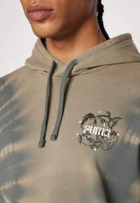 Sweat à capuche tie-dye vert olive en coton, arborant un logo graphique avec une fraise et des feuilles. Fini avec des cordons de serrage gris.