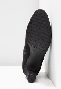 Semelle de chaussure en daim noir affichant le motif du logo "Tamaris" et talon avec le texte "ANTISLIDE", positionnée à la verticale contre un fond d'angle blanc.