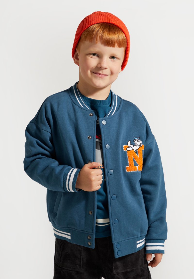 Turkusowa katana uniwersytecka z białymi paskami, z przodu zapinana na guziki, z pomarańczową łatą w kształcie litery 'N'. Nałożona na niebieski sweter.