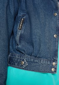 Jeansjacka med en blå-grön tyginredning, prydd med silverknappar, en ficka med en logotypetikett och en något samlad midja.