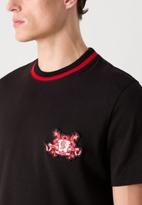 Black cotton t-shirt with a round neck, featuring a textured red and black embroidered logo on the chest and a ribbed red collar.