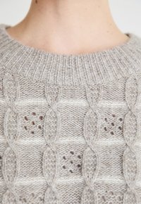 Pull en maille beige clair avec un col côtelé et un motif texturé avec des torsades et de subtils éclats. Tissu doux de poids moyen.